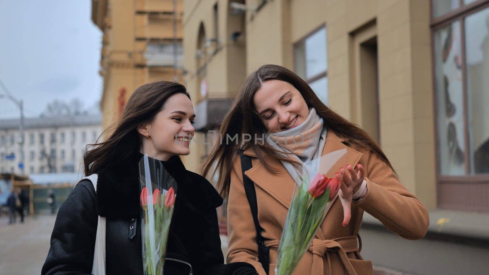 Two young girls, friends with flowers in their hands, walk along a city street on a cloudy spring day.
