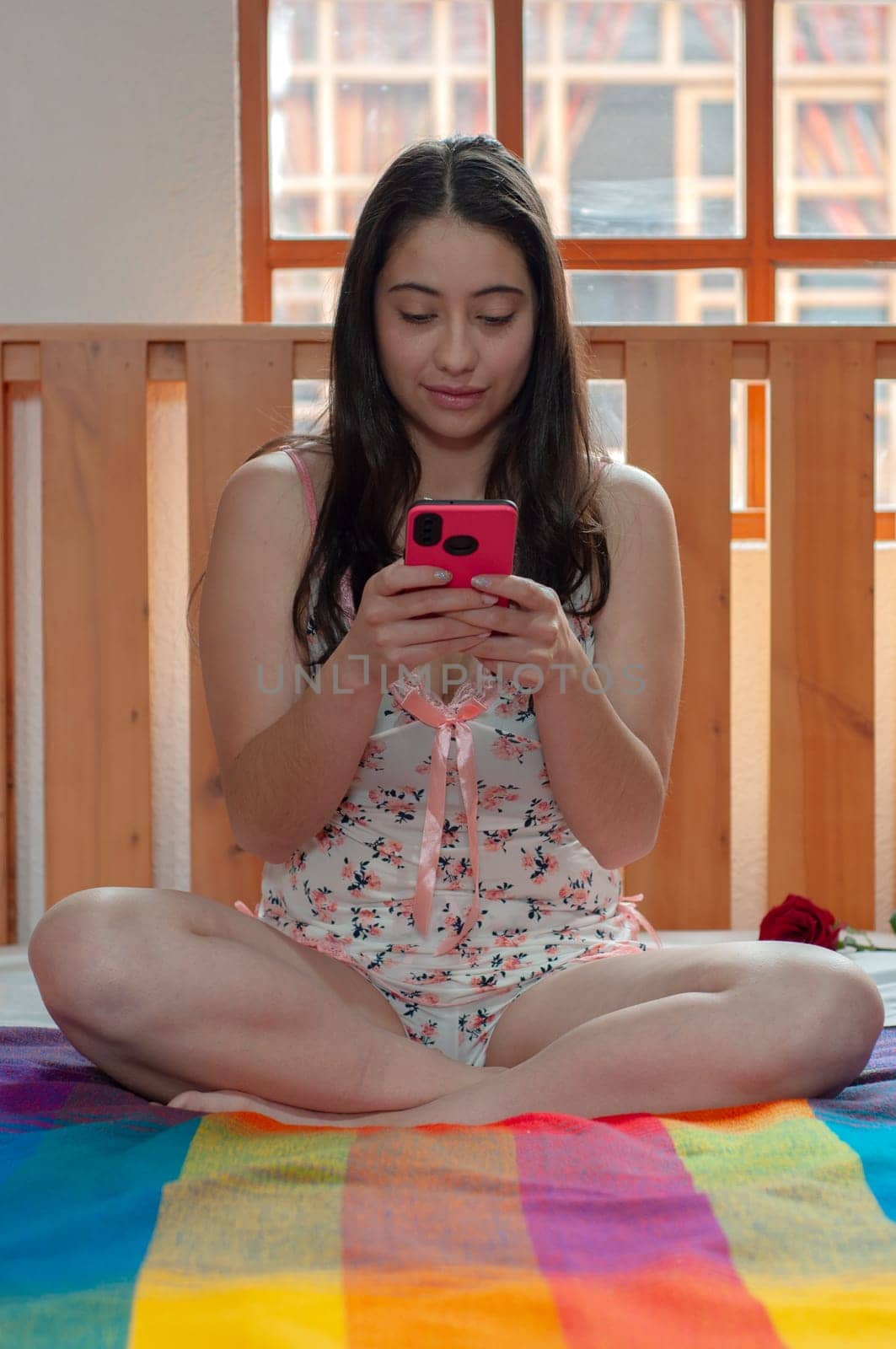 latin american woman writing a message with her cell phone while sitting on her bed in her nightclothes. High quality photo