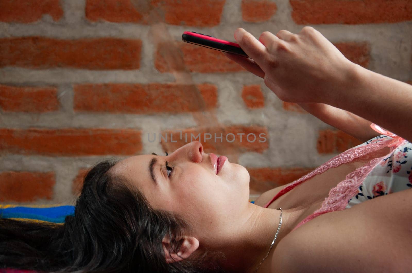 portrait of a girl from venezuela lying face up on her bed typing a text message with a black and pink cell phone by Raulmartin