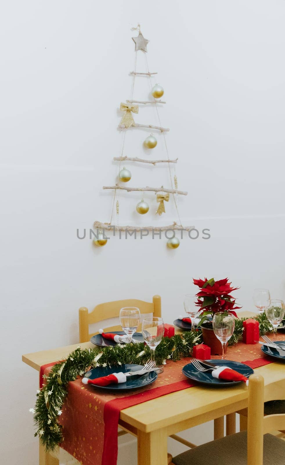 A beautifully served Christmas table with a red tablecloth and a wall-mounted tree made of sticks and branches on a winter evening in the kitchen, close-up side view.