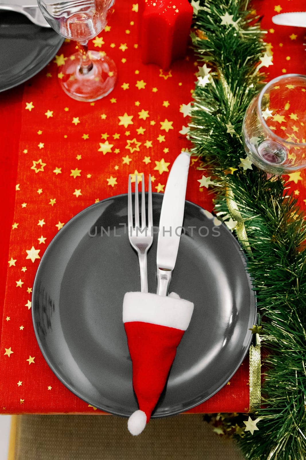 One gray plate with a knife and fork in a mini Santa Claus hat stand on a beautifully served holiday table with a red tablecloth and green tinsel on a winter evening in the kitchen, close-up top view with selective focus.