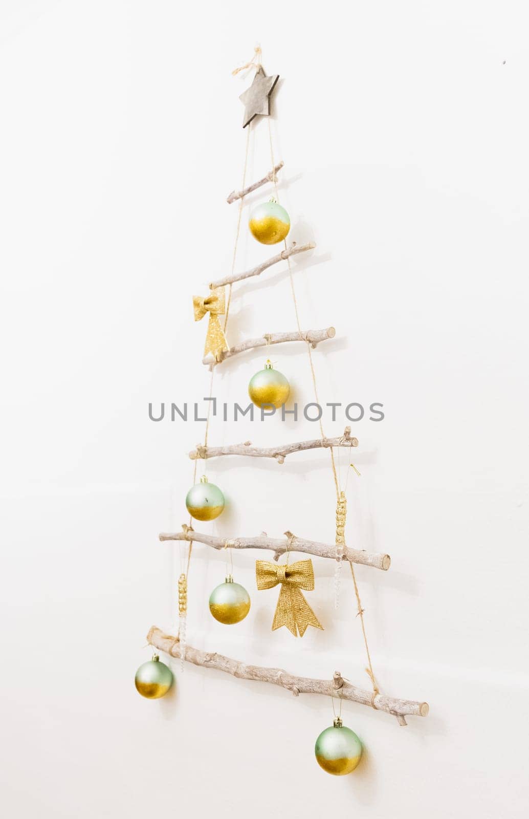 One creative homemade Christmas tree made of branches and jute thread with golden bows and Christmas balls and toys hangs on a white wall on a winter evening in the room, side view close-up.