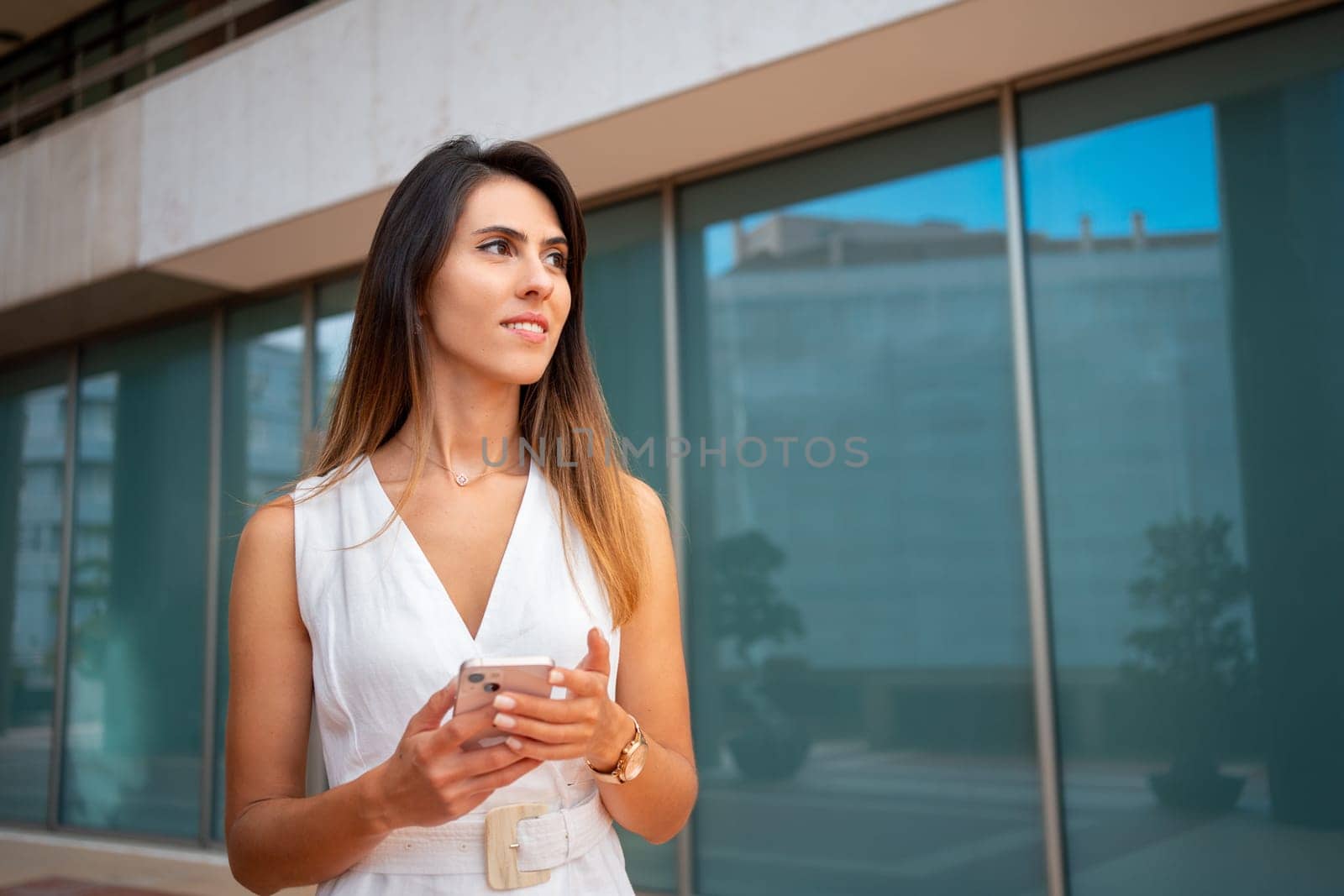 Woman standing outdoor with mobile phone look away. Woman hold smartphone in hand, standing on street outside an office building, waiting for friend. Businesswoman outside