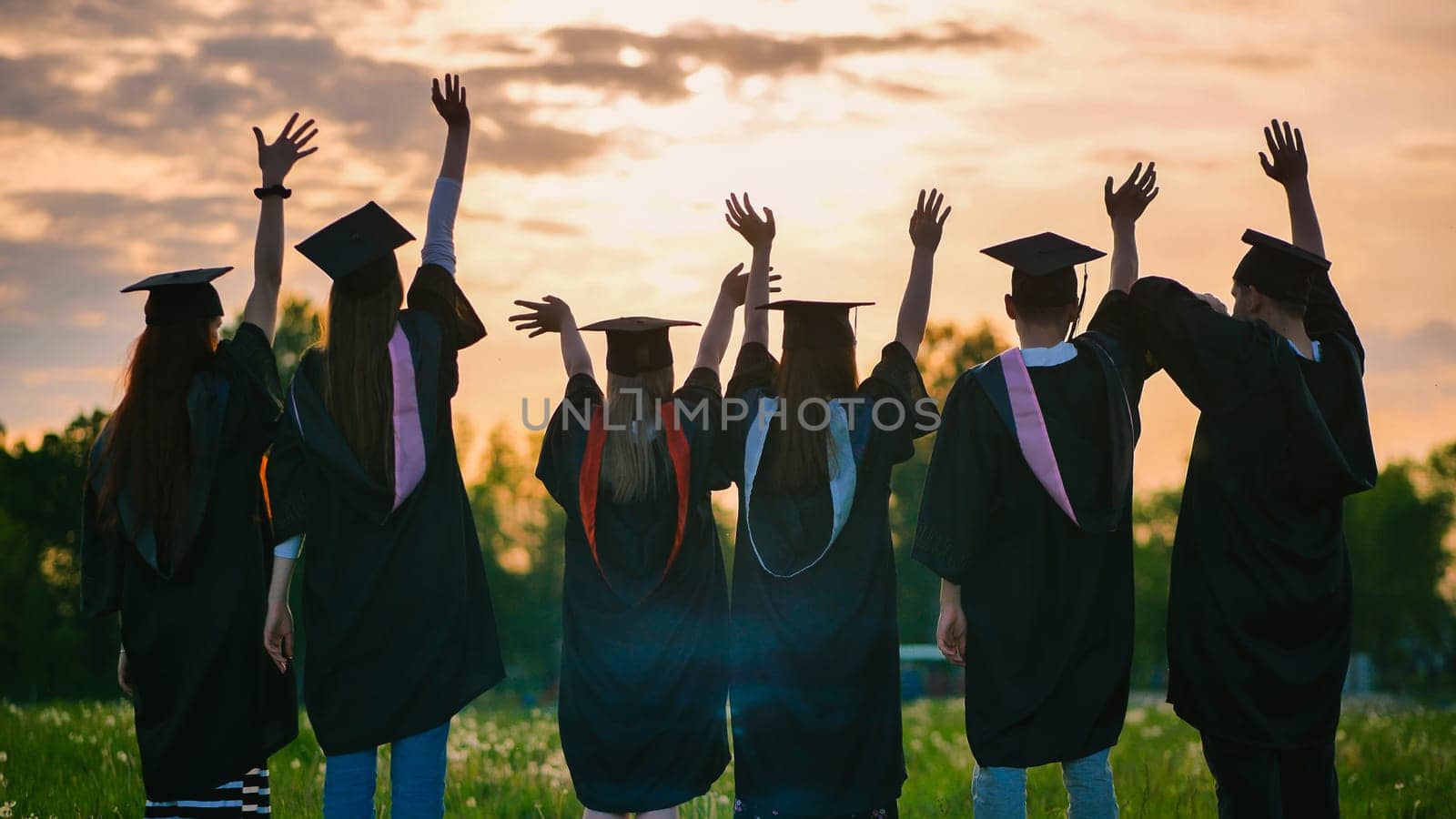 Silhouettes of graduates in black robes waving their arms against the evening sunset