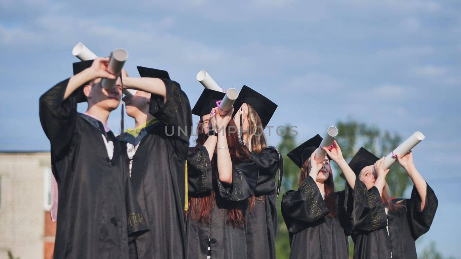 Cheerful graduates on a sunny day look through diplomas like a telescope. by DovidPro