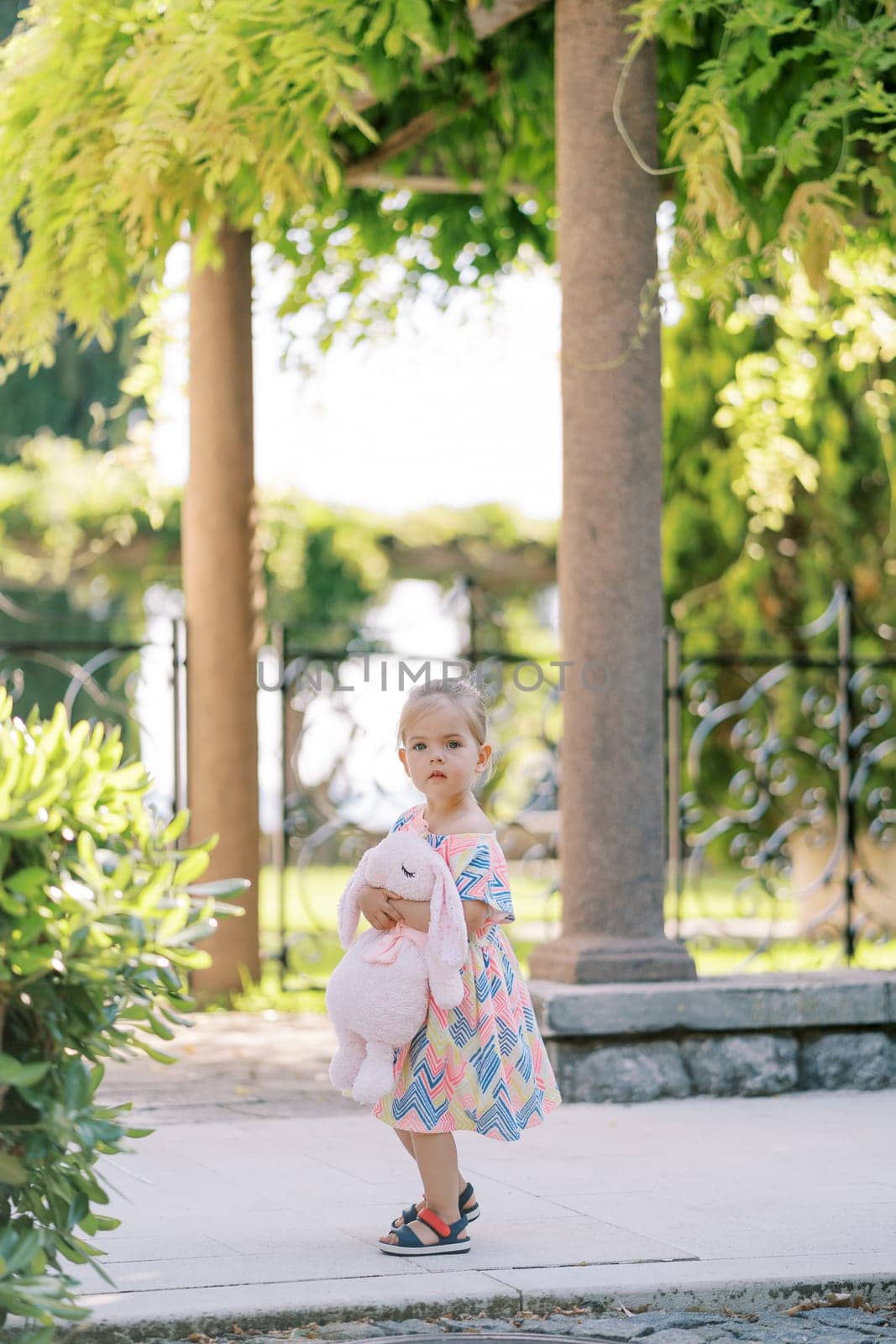 Little girl stands near a pergola column hugging a pink plush hare by Nadtochiy