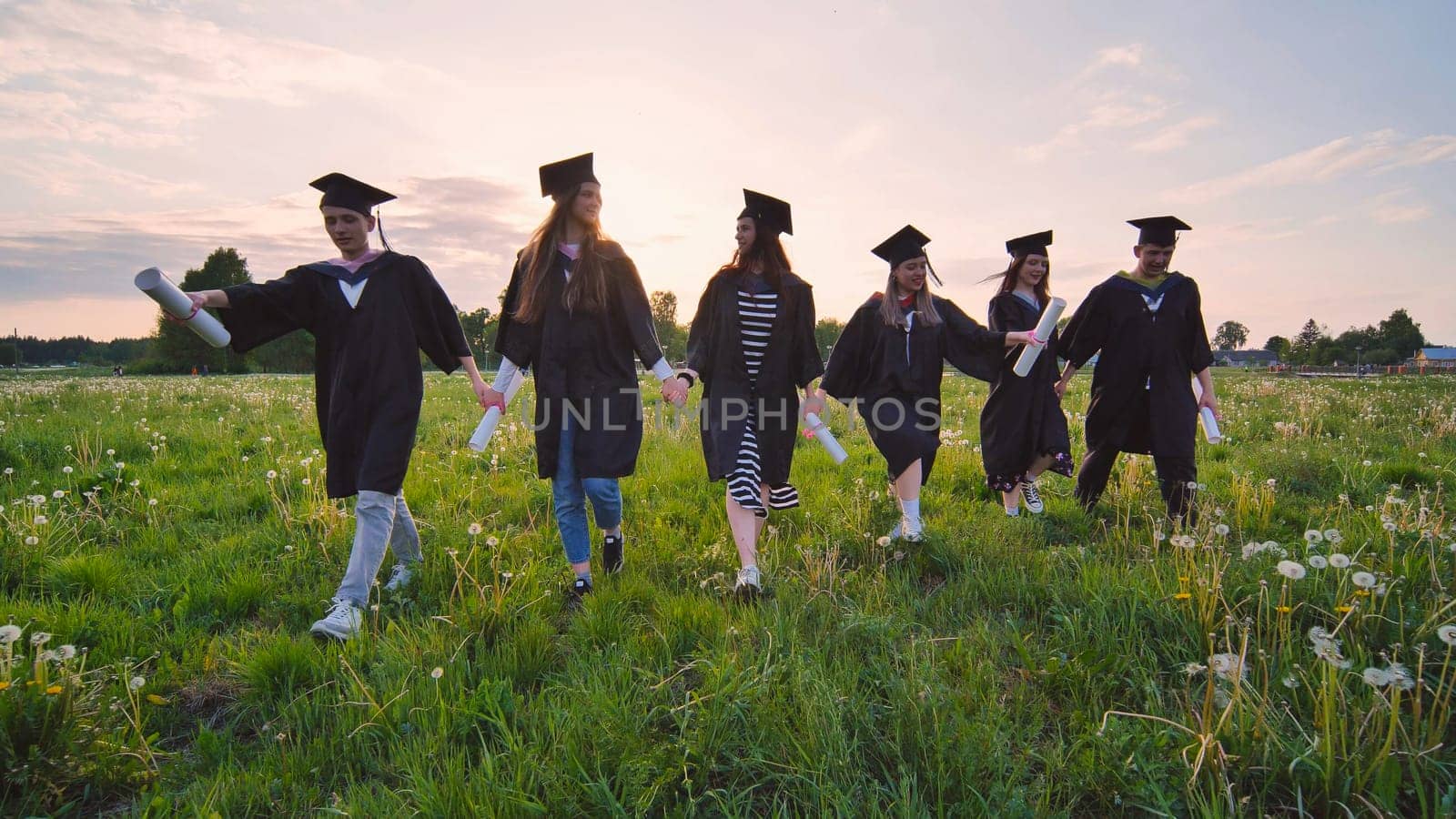 Six graduates in robes walk against the backdrop of the sunset