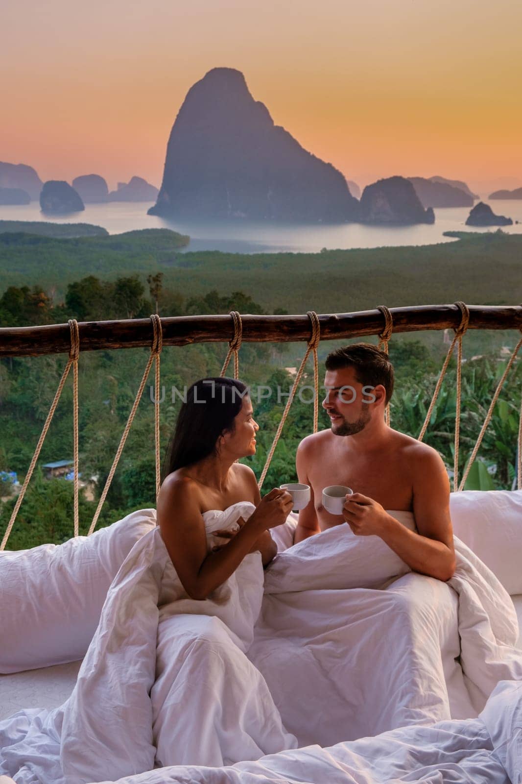 A couple in an outdoor bedroom watching the sunrise over Sametnangshe mountains in Phangnga Bay with mangrove forest in the Andaman Sea, men and woman in an outdoor bed at Phangnga, Thailand