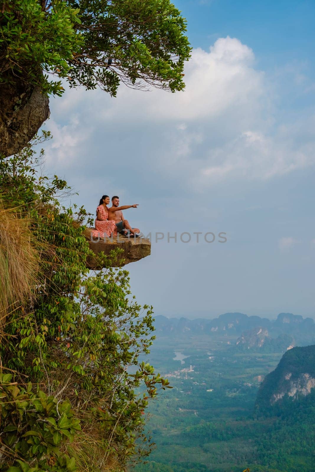 Couple at the edge of a mountain, Dragon Crest Mountain Krabi Thailand, Young travelers sit on a rock that overhangs the abyss, with a beautiful landscape. Dragon Crest or Khuan Sai at Khao Ngon Nak