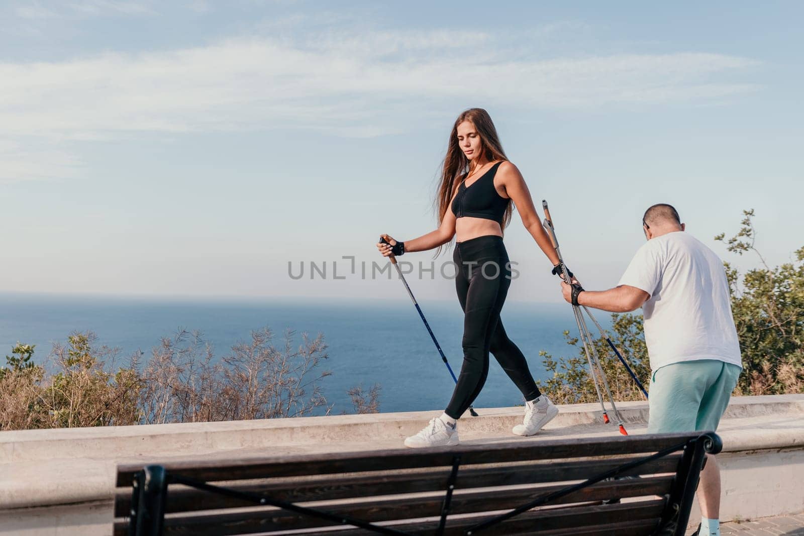 Happy Middle aged couple or friends practicing nordic walking in park near sea. Mature couple with trekking poles walking, practicing Nordic walking outdoors. Aging youthfully and sport concept by panophotograph