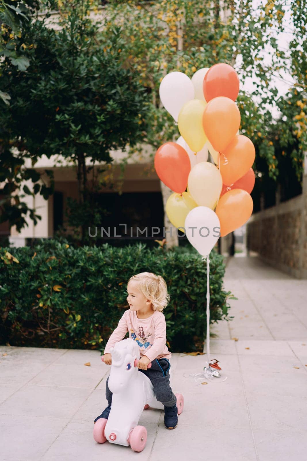 Little girl rides a unicorn balance bike in the garden against the background of colored balloons. High quality photo