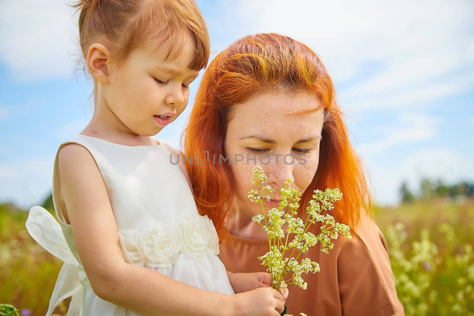 Happy female family with mother and daughter on green and yellow meadow full of grass and flower. Woman with red hair and blonde girl having fun, joy and hug in sunny summer day. Concept family love
