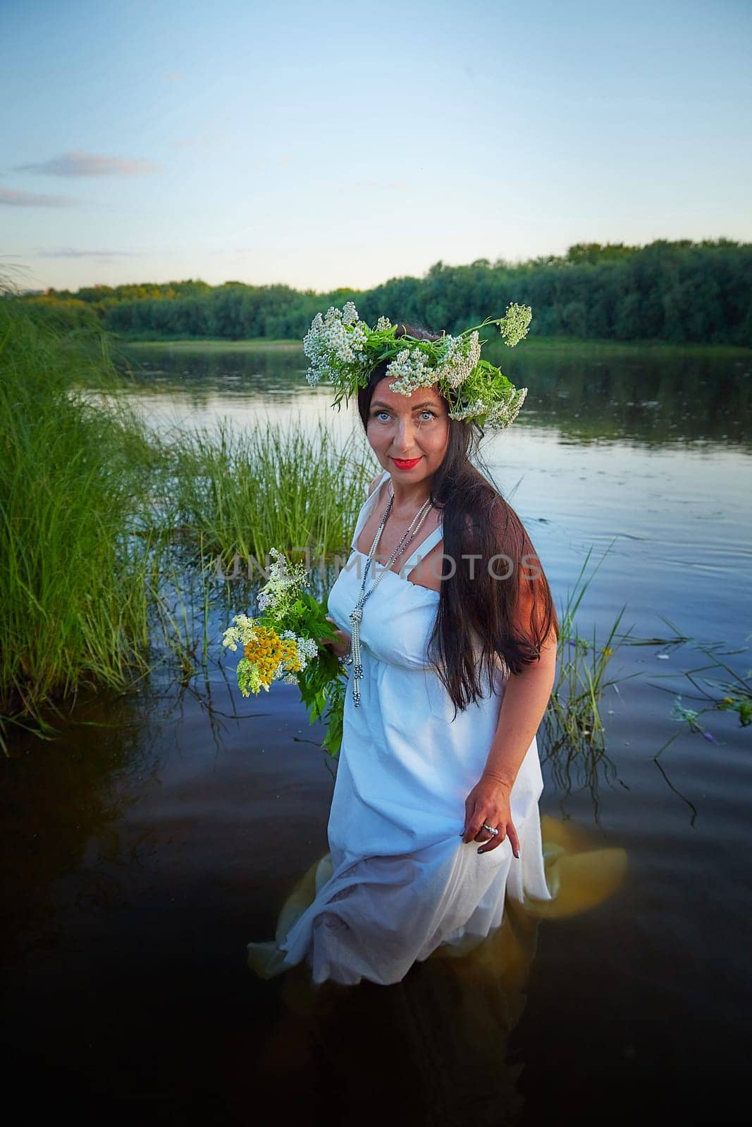 Adult mature brunette woman in a white dress, sundress and a wreath of flowers in summer in water of river or lake in the evening at sunset. Celebration of the Slavic pagan holiday of Ivan Kupala by keleny