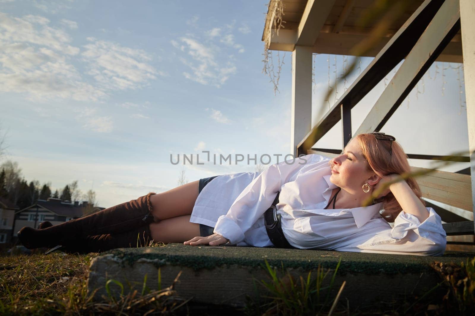 Beautiful girl with red hair in white shirt in open wooden pavillion in village or small town. Young slender woman and sky with clouds on background on an autumn, spring or summer day by keleny