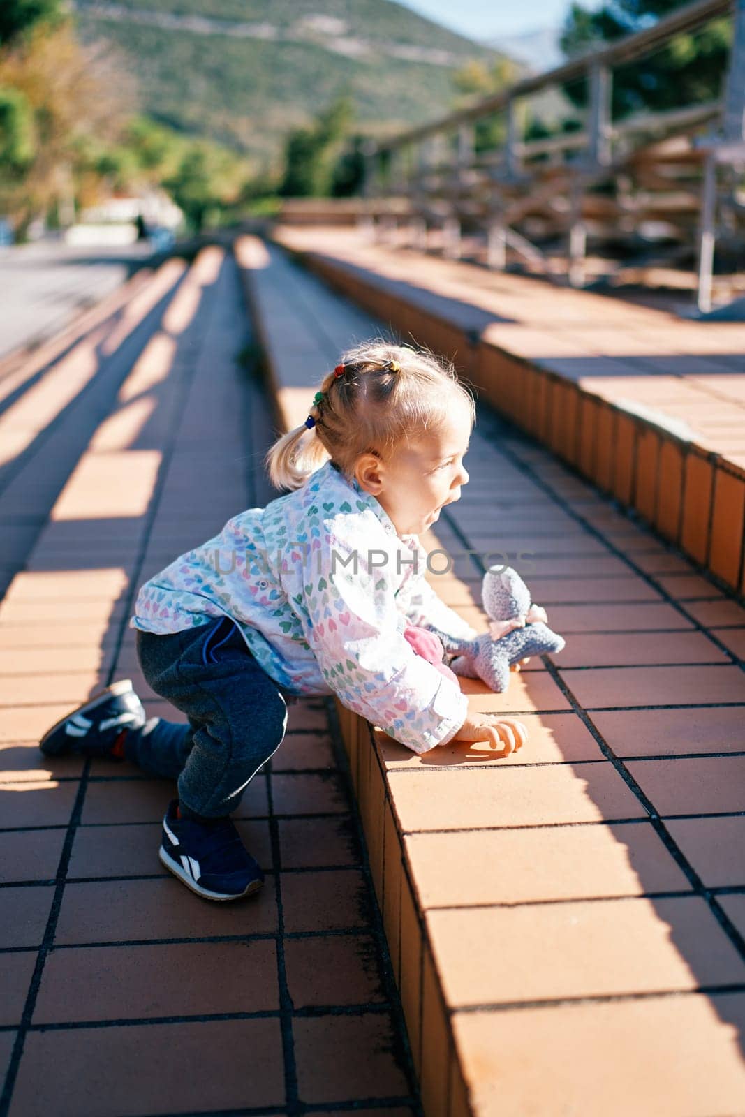 Little girl climbs the steps in the park on all fours with a toy in her hand by Nadtochiy