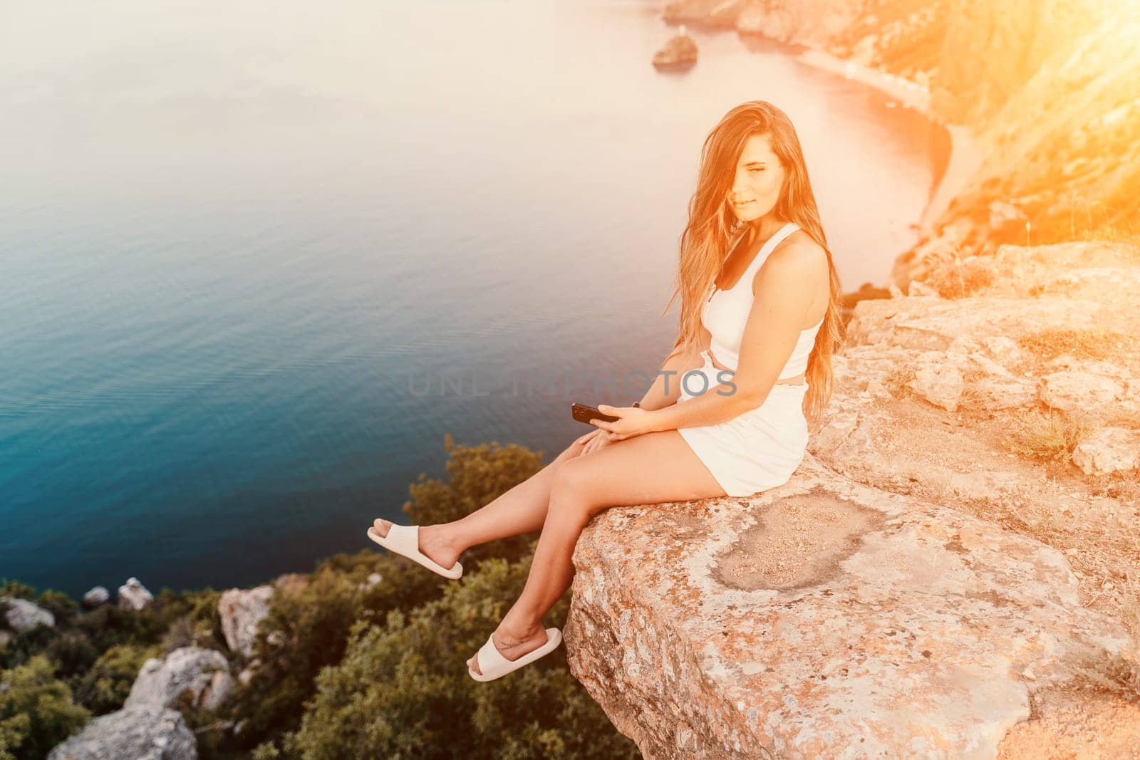 Happy woman in white shorts and T-shirt, with long hair, talking on the phone while enjoying the scenic view of the sea in the background. by Matiunina