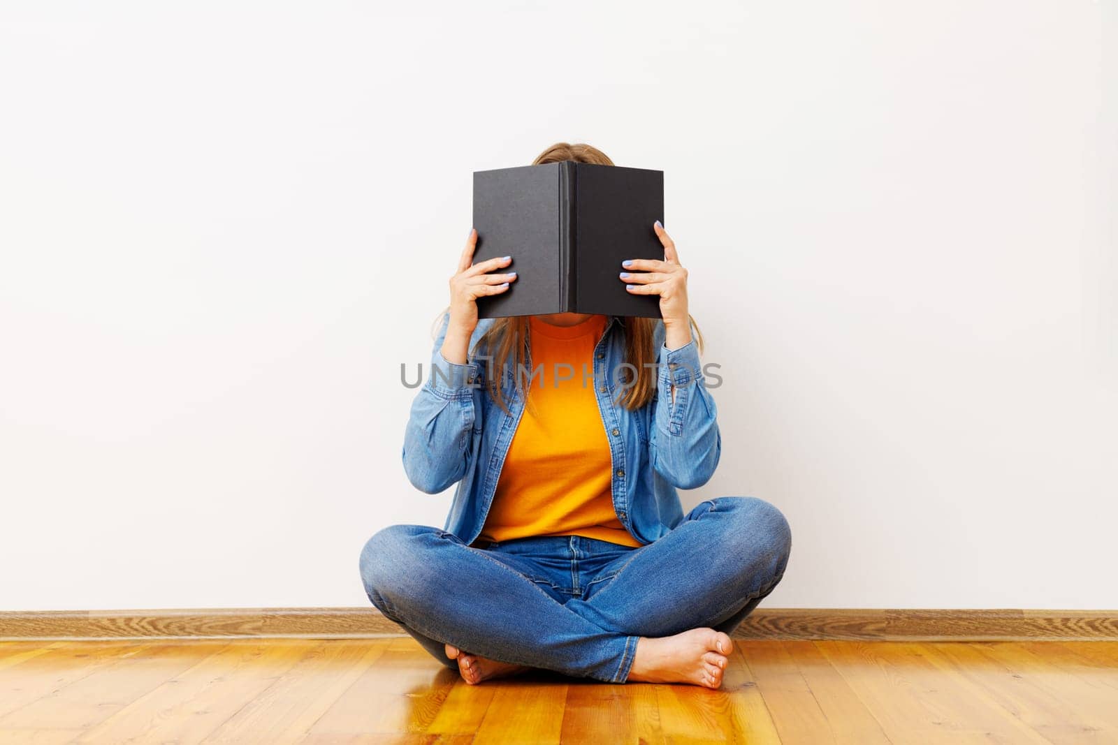 A young woman covers her face with a book as she sits on floor against the wall by andreyz