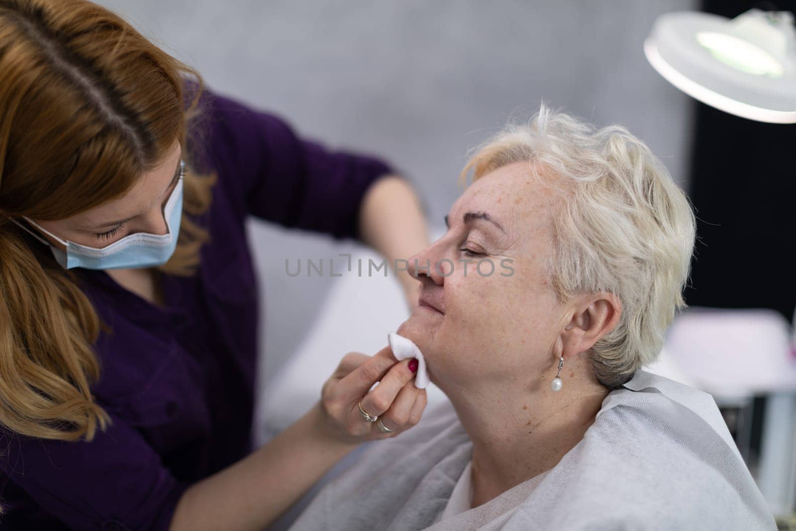 A client of a beauty salon is sitting in the salon. The beautician wipes her client's face with a cosmetic swab.
