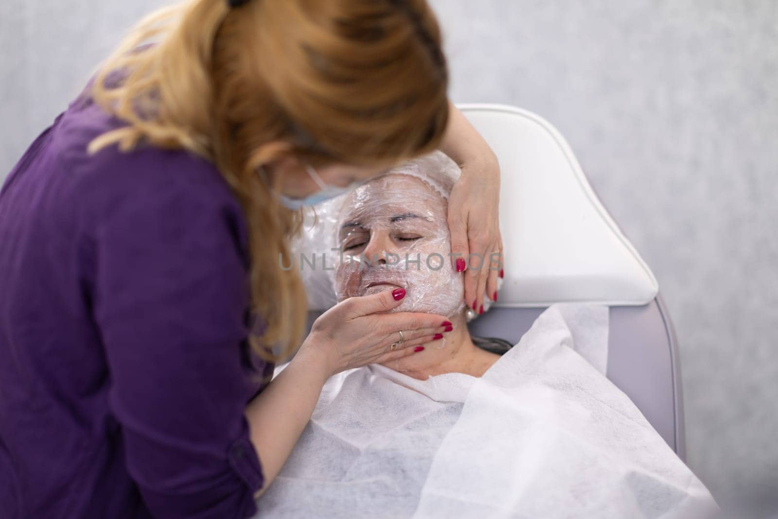 An elderly woman sits in an armchair. Her body is covered with a white covering. On her head is a disposable sanitary cap. The beautician is performing a facial treatment for the client.