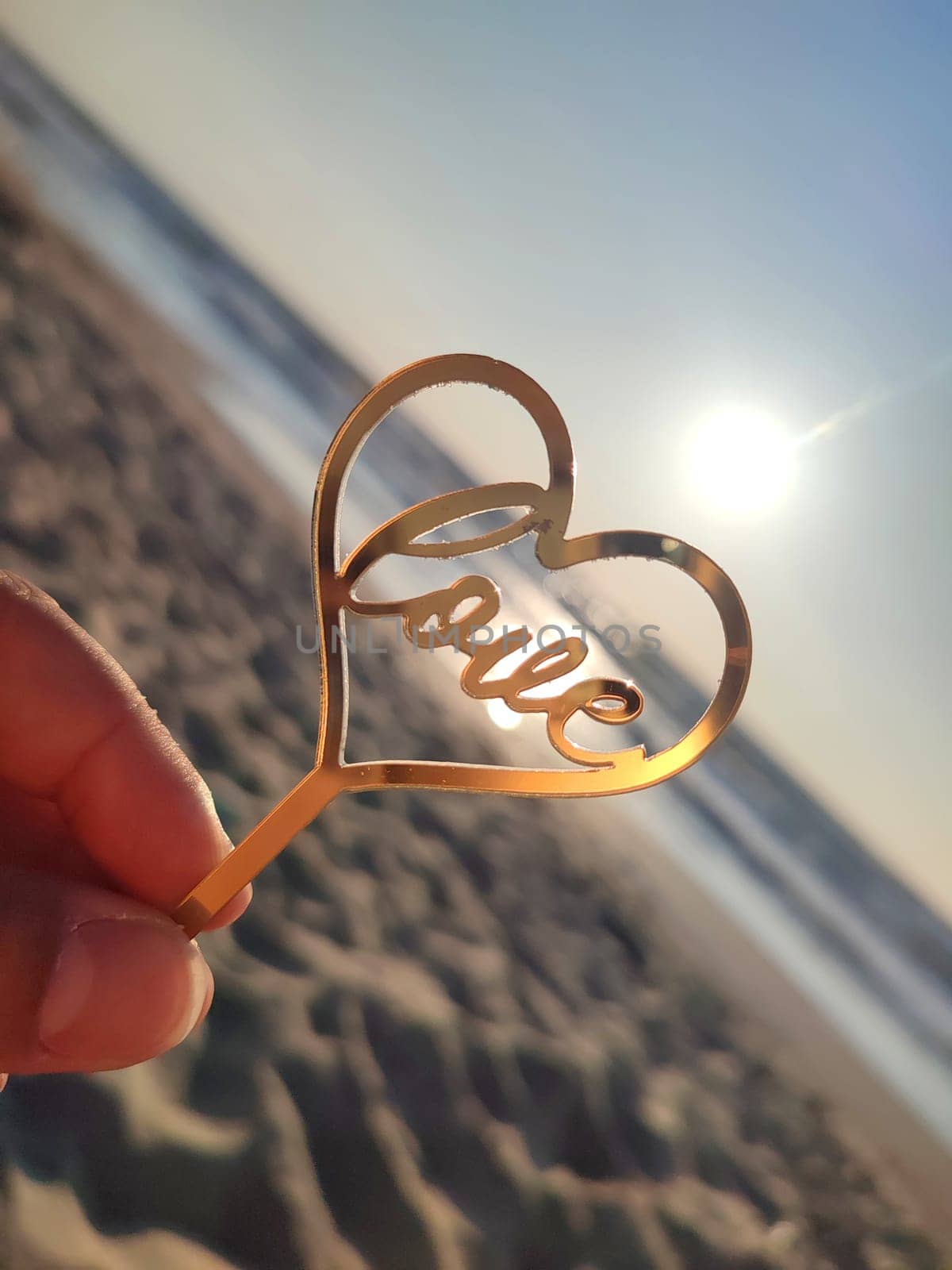 Person holding in fingers hand stick in shape golden heart and word Love on background sea and sea waves in seashore on sunny summer day close-up. Concept love romance amour St Valentines Day