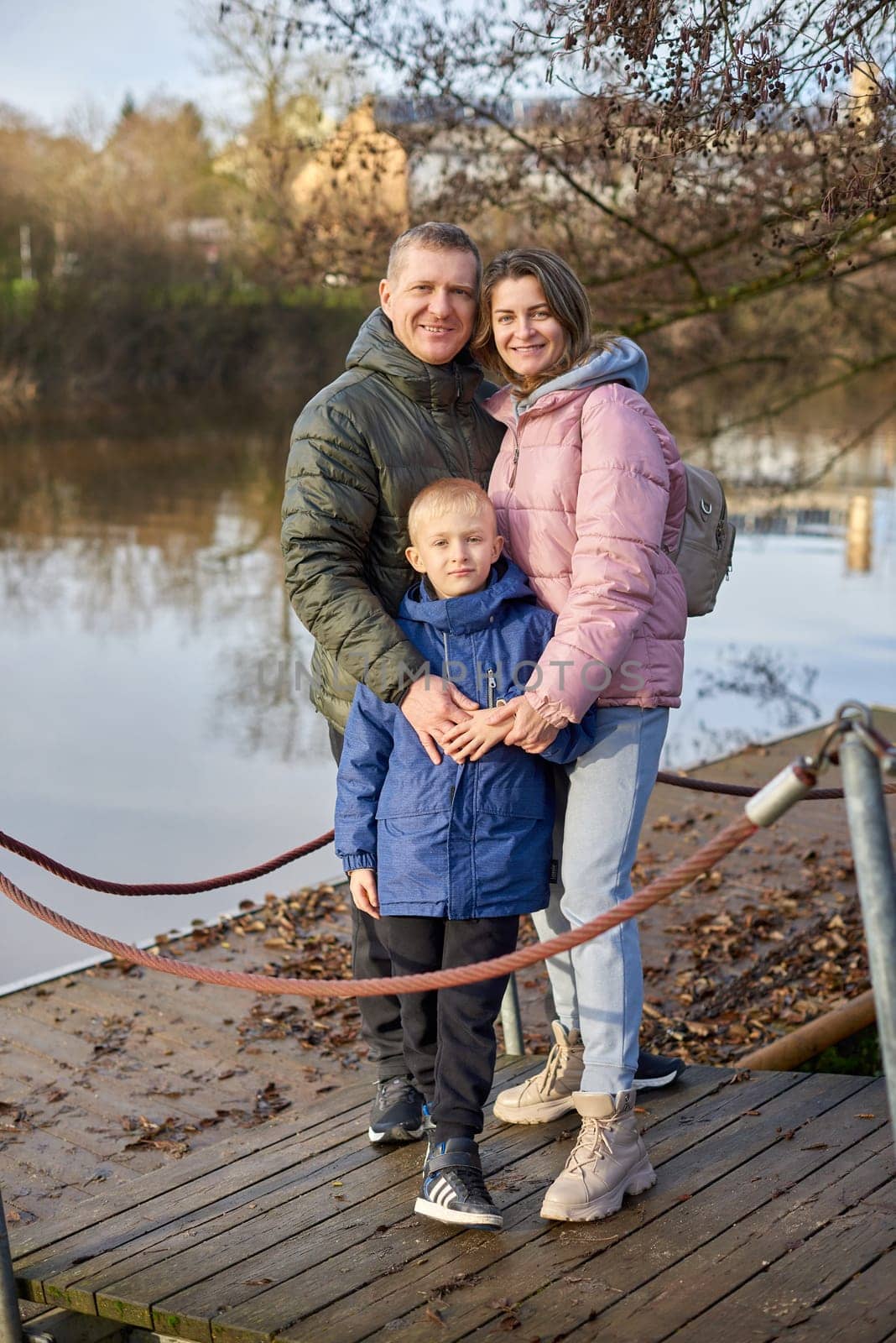 Young parents and their little son standing on the pier near the lake or river shore, at sunset on autumn day. Family Harmony by the Autumn Shore: Standing Strong Together. A Day of Family Love: Autumn Serenity by the Standing Lake by Andrii_Ko