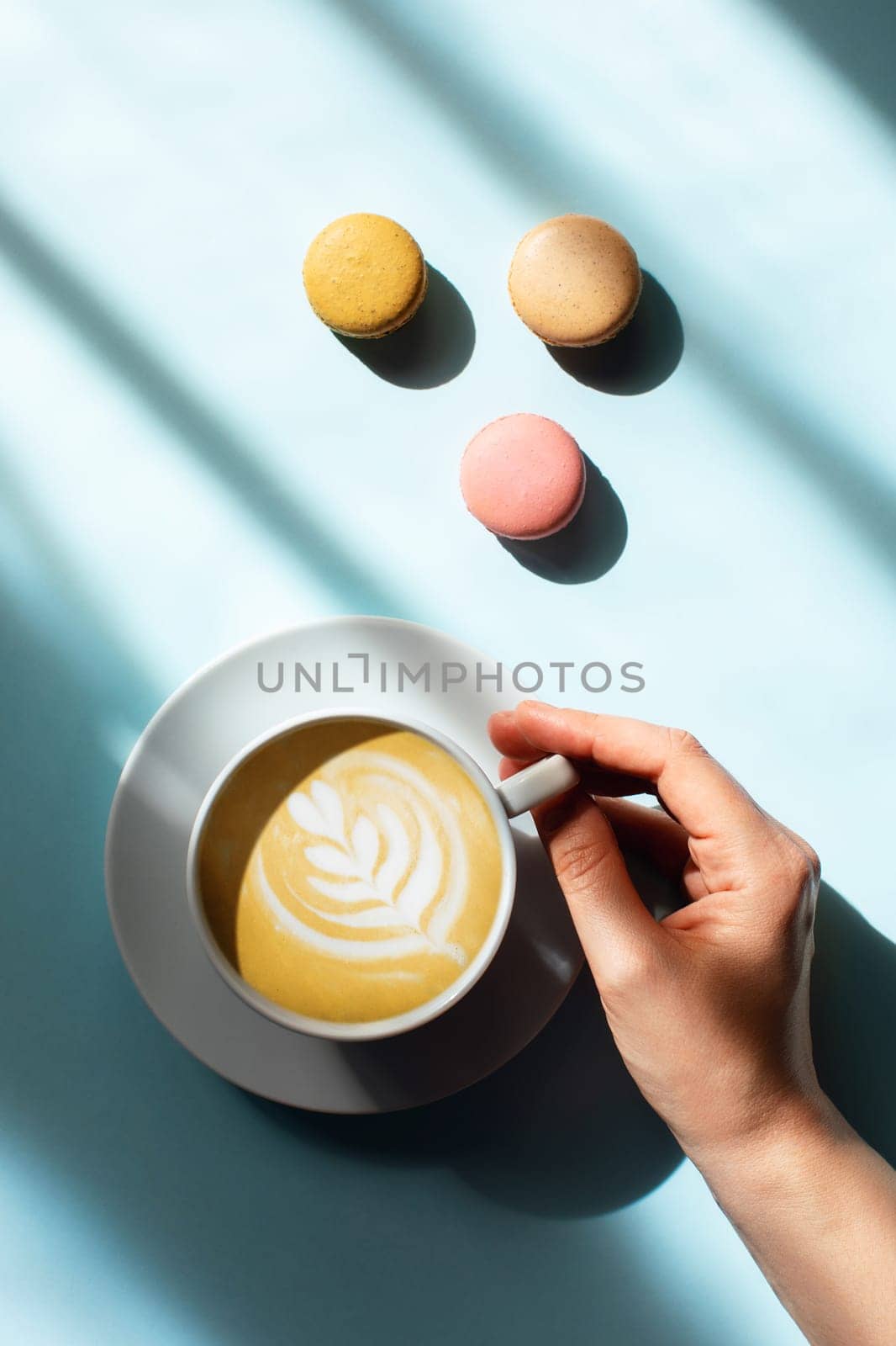 Blue background with cup of tasty coffee in a woman hand and color macaroons in sunlight.