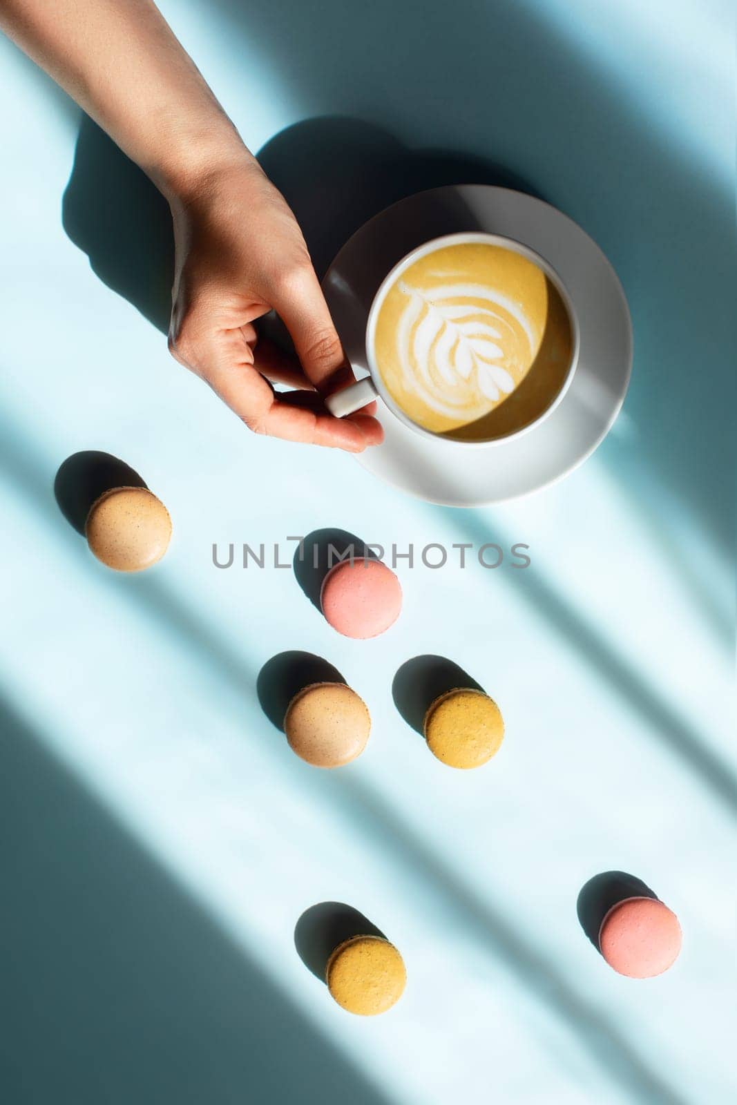 Blue background with cup of tasty coffee in a woman hand and color macaroons in sunlight near the window.