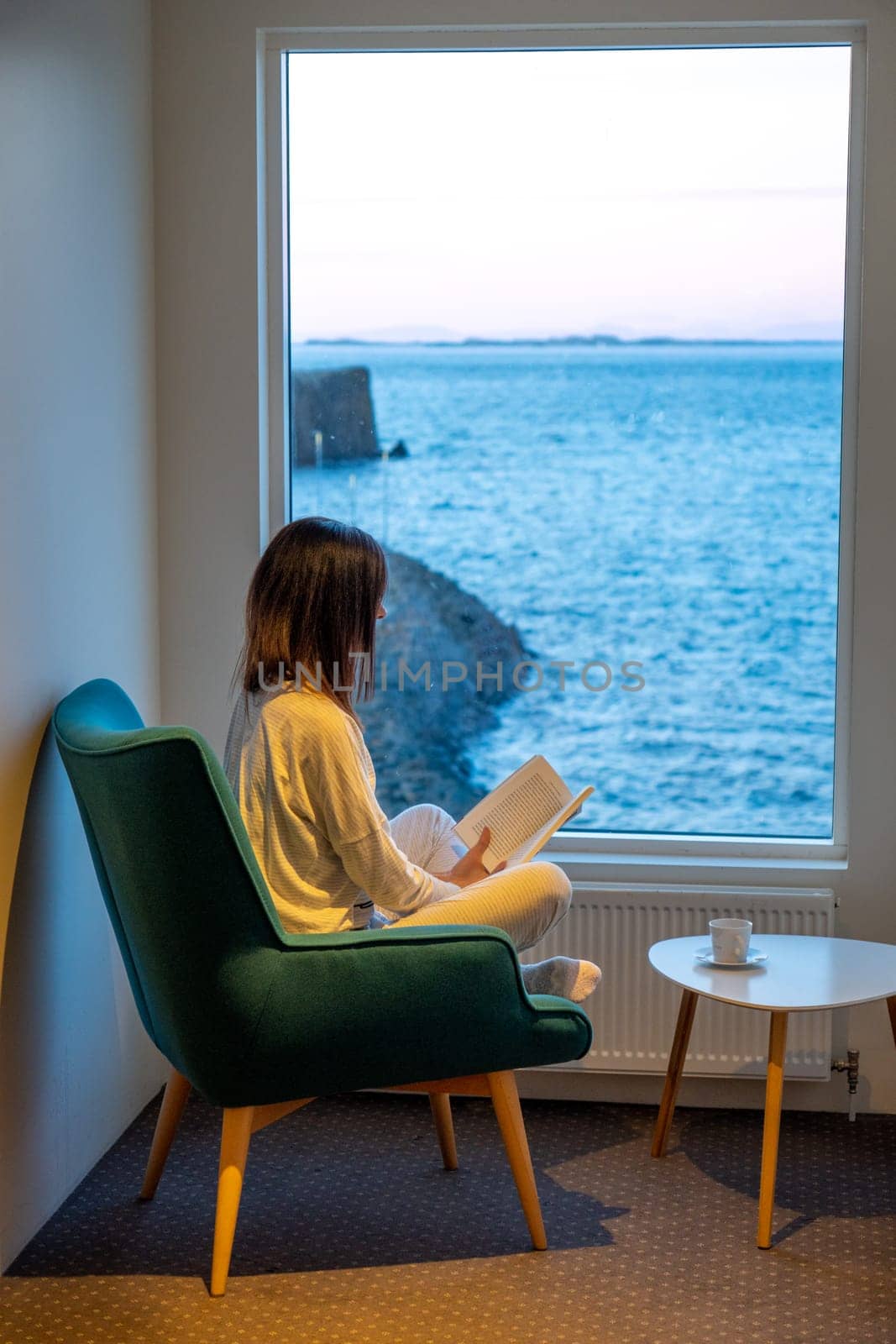 Woman in pajamas reading in a window facing the sea at sunset