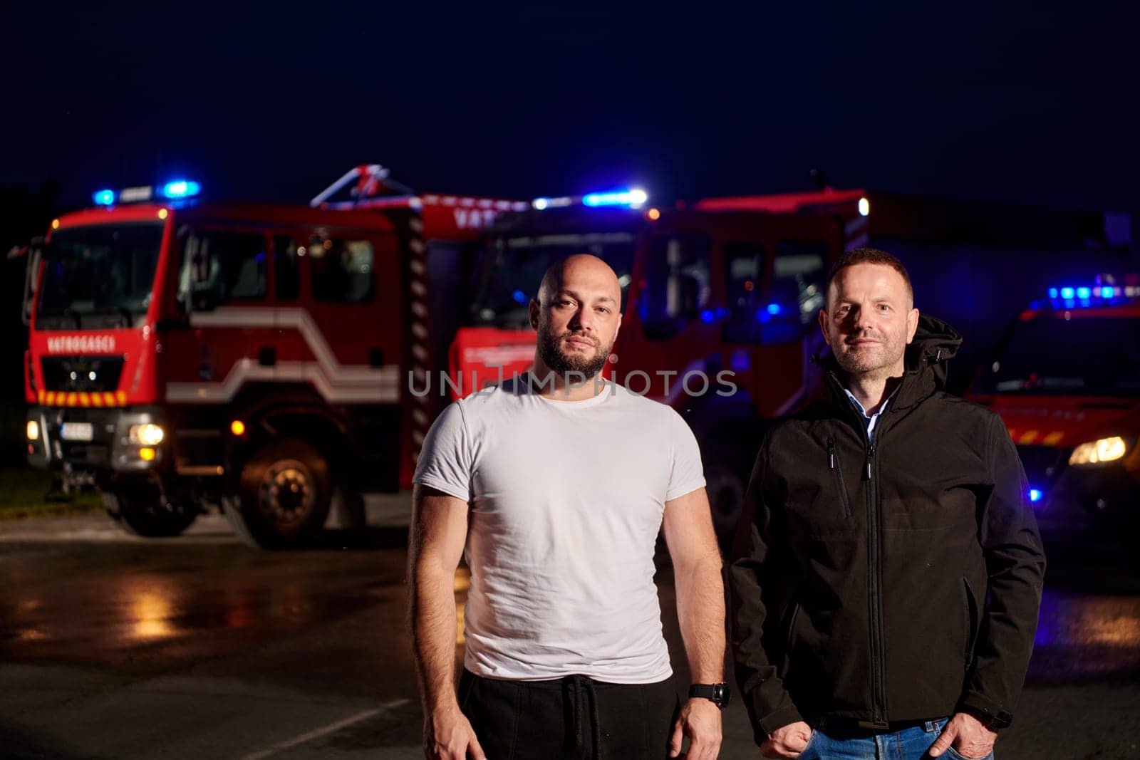 Group of firefighters, dressed in civilian clothing, stand in front of fire trucks during the night, showcasing a moment of camaraderie and unity among the team as they reflect on their duties and the challenges faced during their firefighting service.