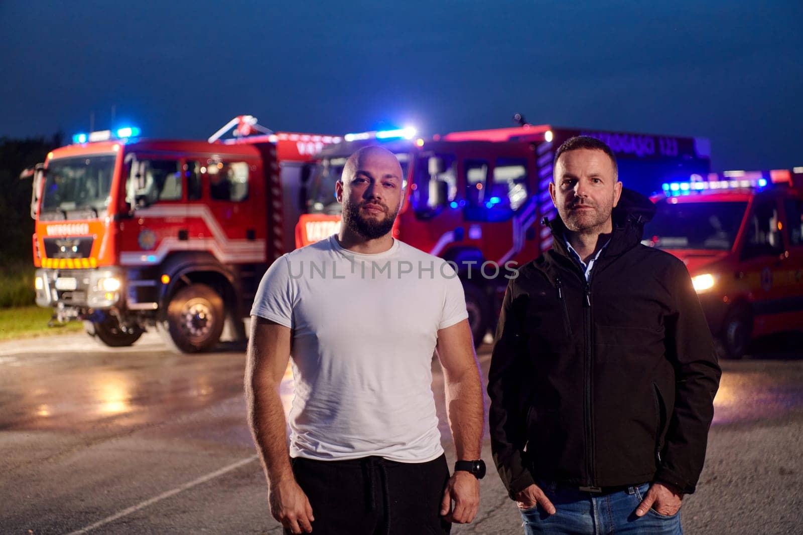 Group of firefighters, dressed in civilian clothing, stand in front of fire trucks during the night, showcasing a moment of camaraderie and unity among the team as they reflect on their duties and the challenges faced during their firefighting service by dotshock