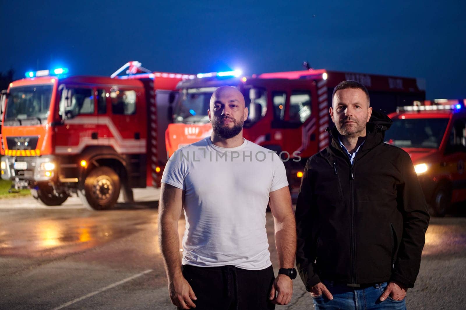 Group of firefighters, dressed in civilian clothing, stand in front of fire trucks during the night, showcasing a moment of camaraderie and unity among the team as they reflect on their duties and the challenges faced during their firefighting service.