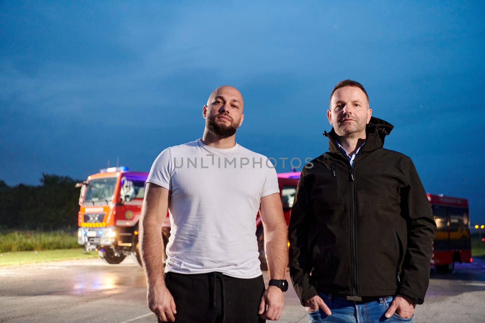 Group of firefighters, dressed in civilian clothing, stand in front of fire trucks during the night, showcasing a moment of camaraderie and unity among the team as they reflect on their duties and the challenges faced during their firefighting service by dotshock