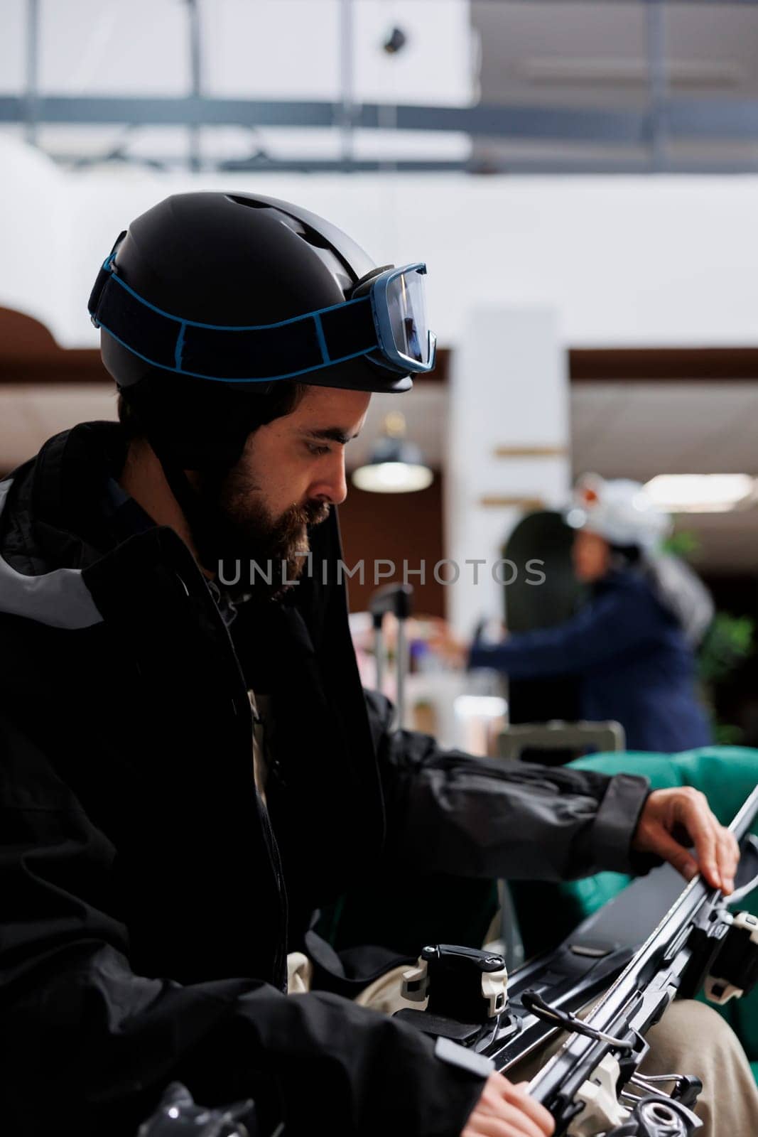 Close-up of caucasian traveler adjusting ski gear in hotel lobby. Male tourist, wearing winter jacket and ski goggles, ensuring safety of wintersports equipment. Ready to explore ski resort for skiing.