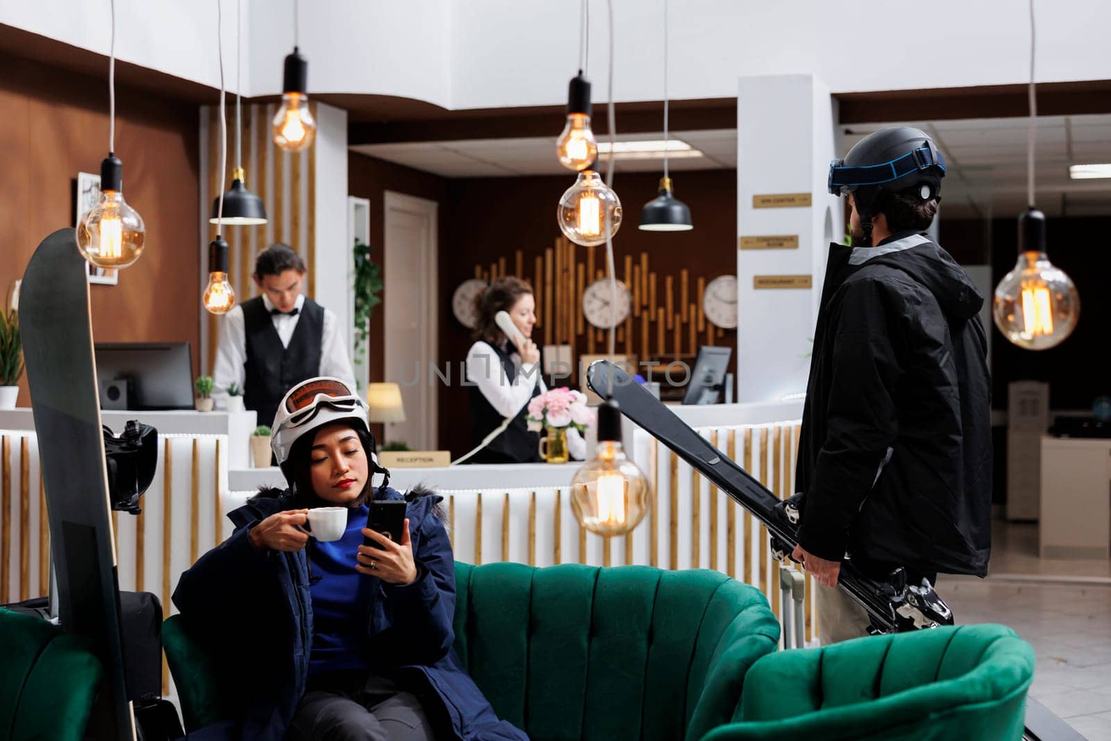 Male traveler with skiing skis arriving at hotel lobby during winter holiday while woman with cup in hand, comfortably seated on sofa browsing mobile device. Receptionists working in the background.
