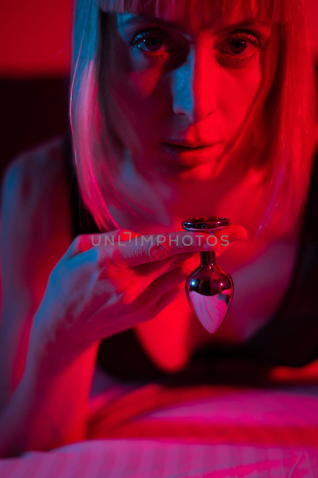 The blonde lies on the bed in red blue neon light and holds a steel butt plug