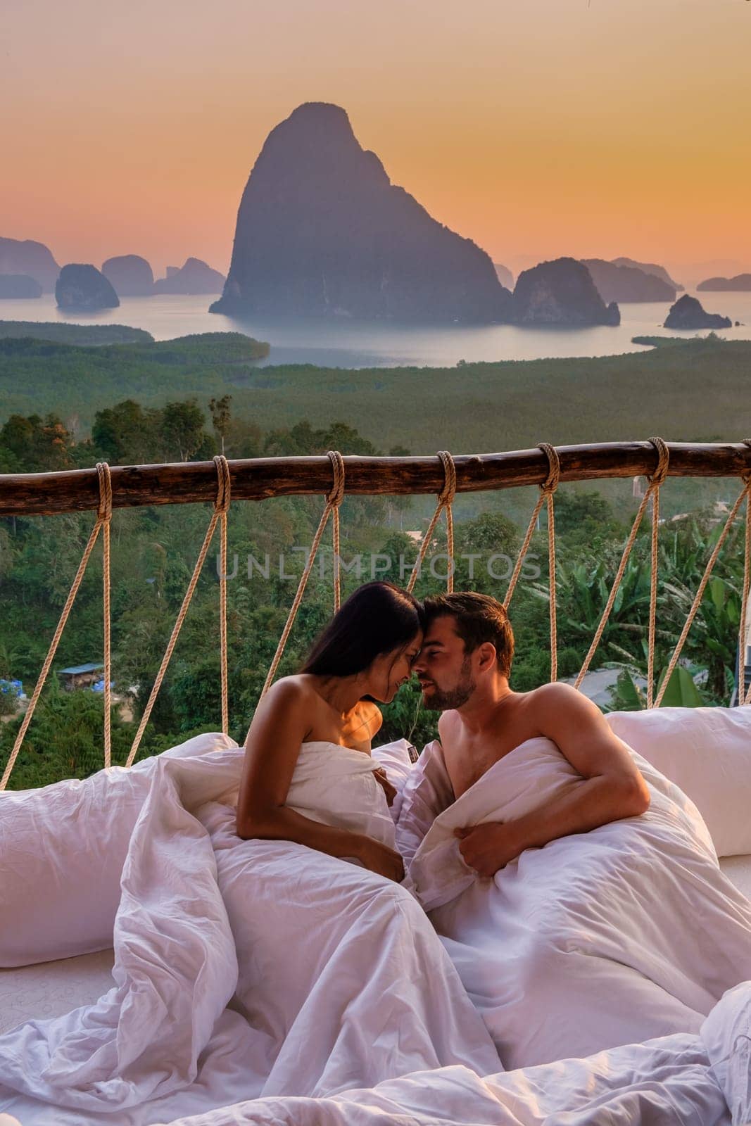 A couple in an outdoor bedroom watching the sunrise over Sametnangshe mountains in Phangnga Bay with mangrove forest in the Andaman Sea, men and woman in an outdoor bed hugging together