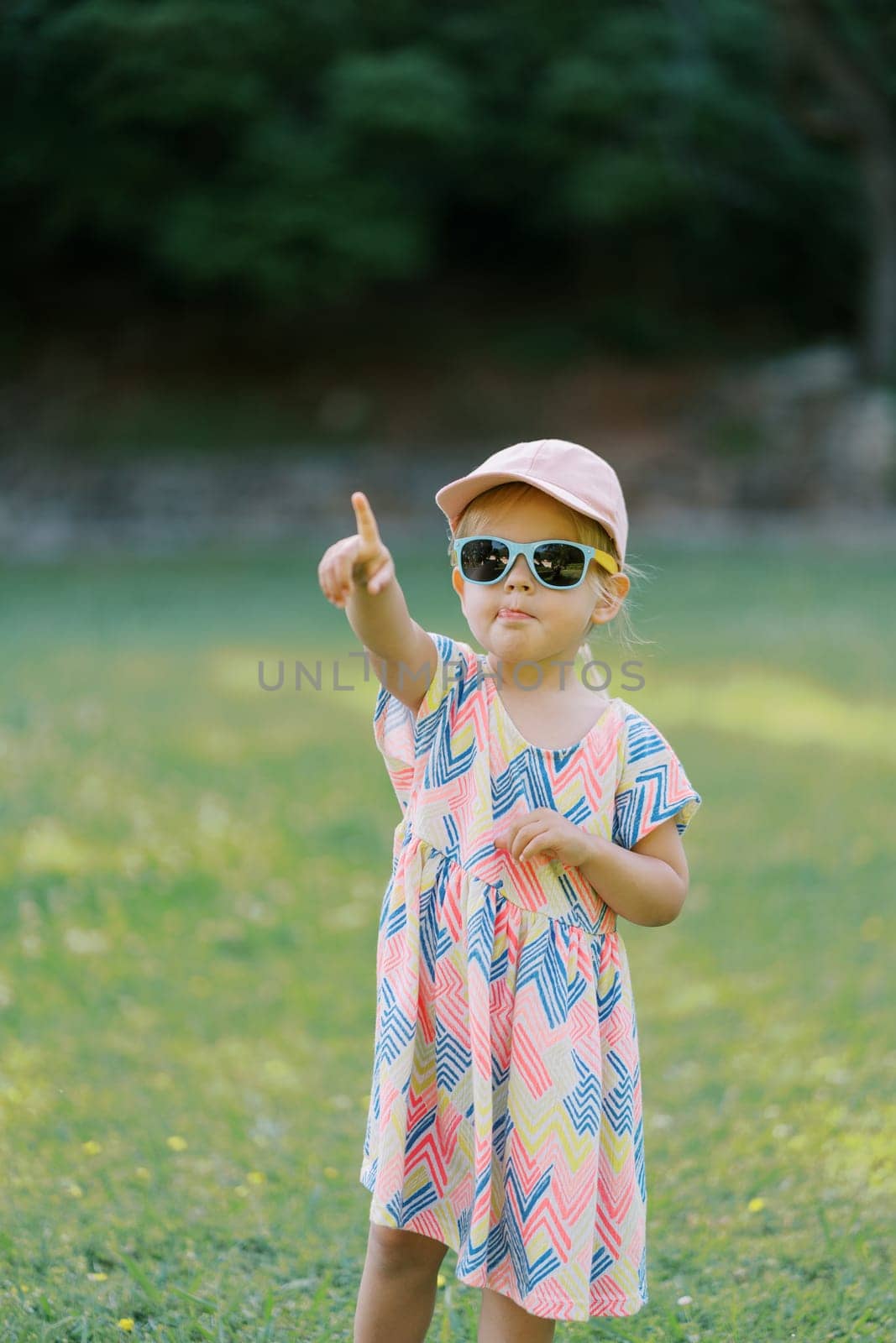 Little girl in sunglasses stands in a clearing pointing her finger into the distance by Nadtochiy