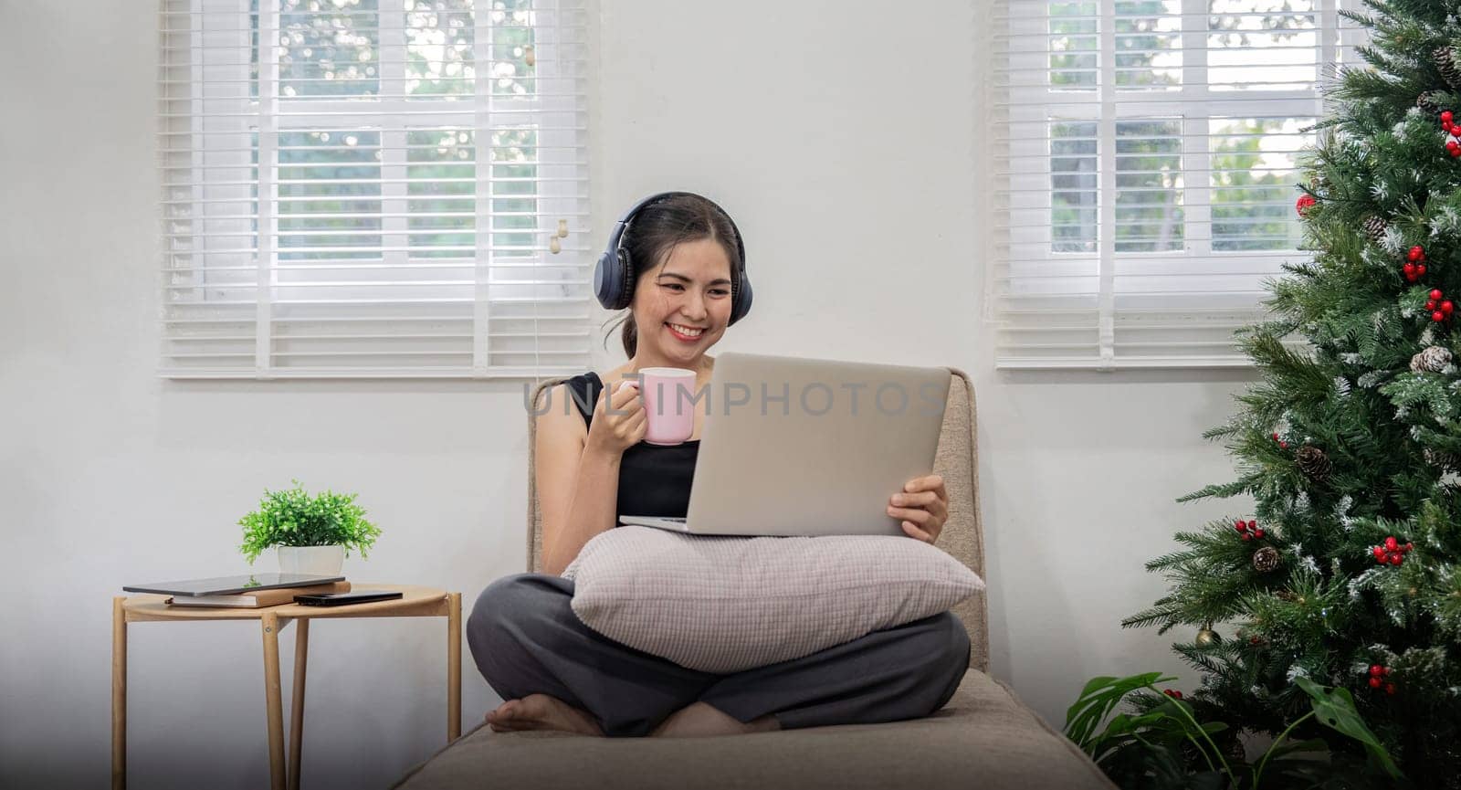 Pretty young woman asian listening to music with laptop relaxing and drinking tea or coffee while sitting on sofa at home by nateemee