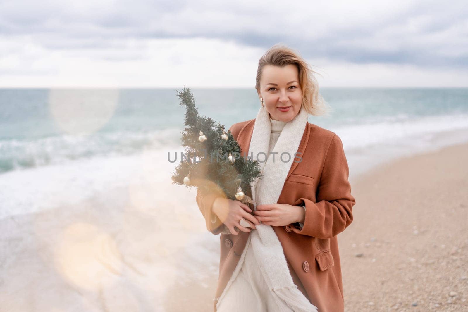Blond woman Christmas sea. Christmas portrait of a happy woman walking along the beach and holding a Christmas tree in her hands. She is wearing a brown coat and a white suit