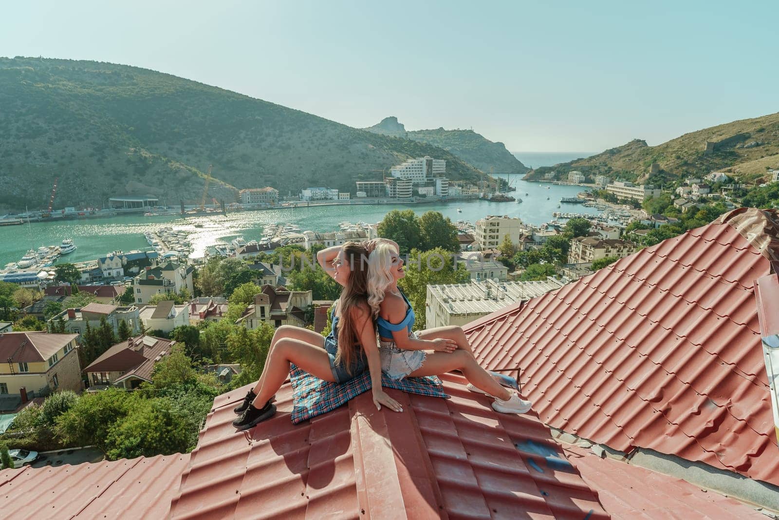 Two women sitting on a red roof, enjoying the view of the town and the sea. Rooftop vantage point. In the background, there are several boats visible on the water, adding to the picturesque scene. by Matiunina