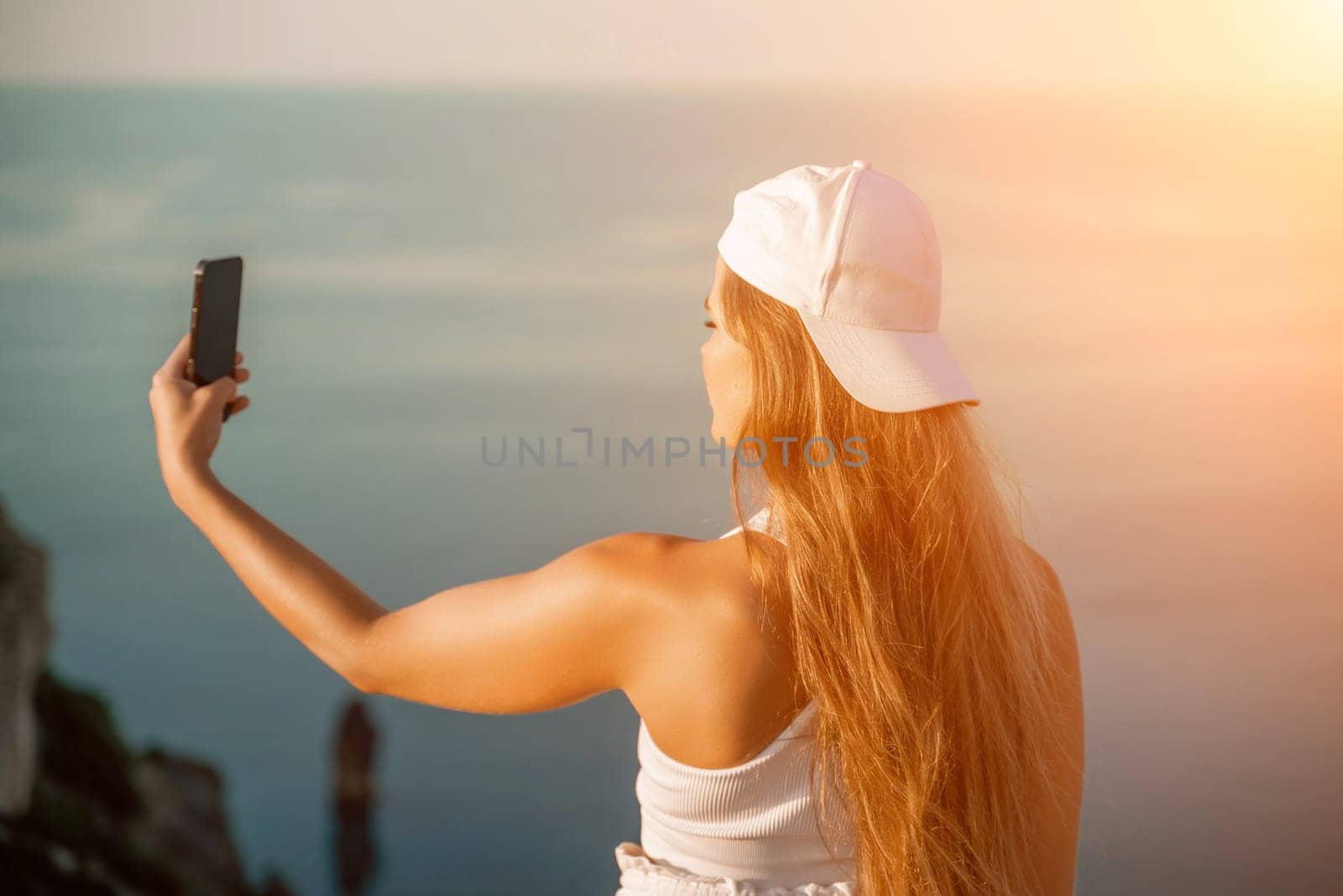 Selfie woman sea. The picture depicts a woman in a cap and tank top, taking a selfie shot with her mobile phone, showcasing her happy and carefree vacation mood against the beautiful sea background by Matiunina