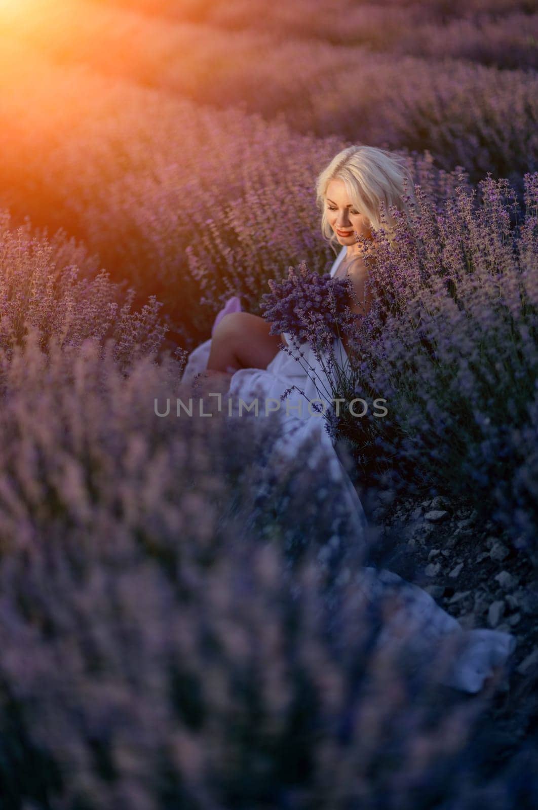 Blonde woman poses in lavender field at sunset. Happy woman in white dress holds lavender bouquet. Aromatherapy concept, lavender oil, photo session in lavender by Matiunina