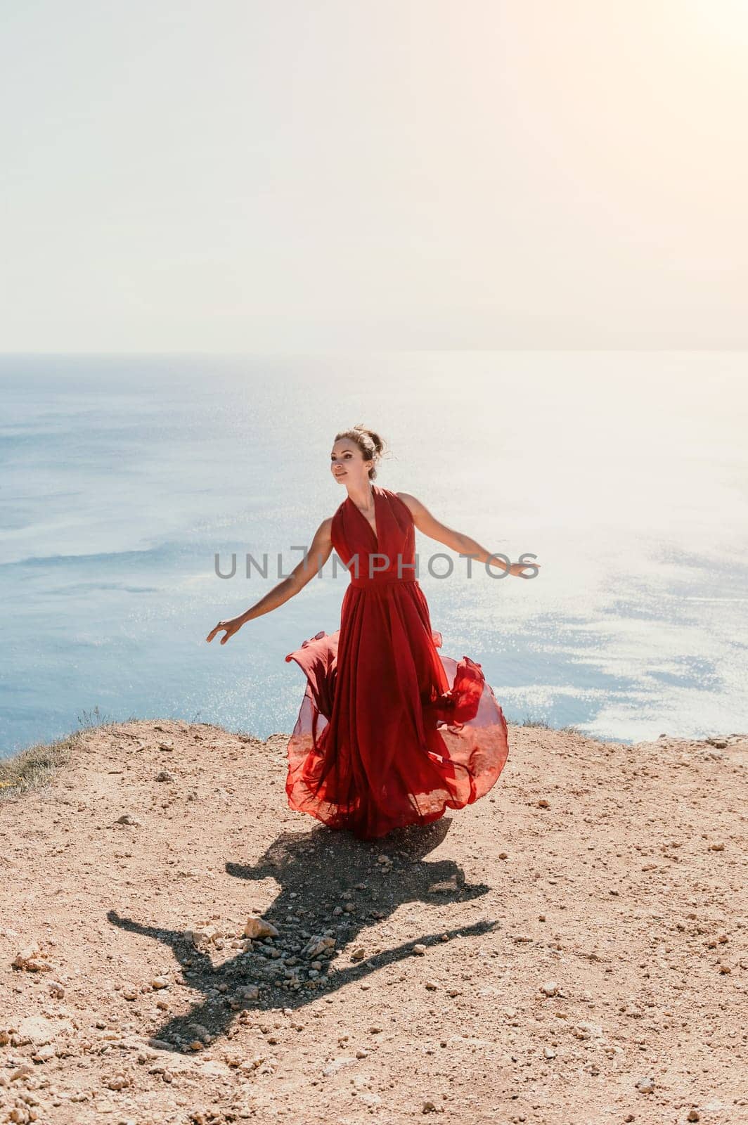 Woman in red dress on sea. Side view a Young beautiful sensual woman in a red long dress posing on a rock high above the sea on sunset. Girl on the nature on blue sky background. Fashion photo. by panophotograph