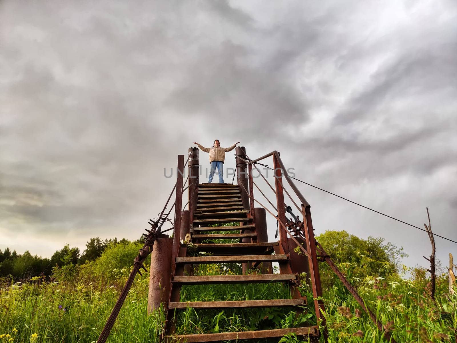 High, old, rusty iron staircase and tiny statue of girl or woman at the top against backdrop of gray, gloomy, thunderous clouds. Concept of an unusual, fantastic journey, surrealism, a strange world by keleny