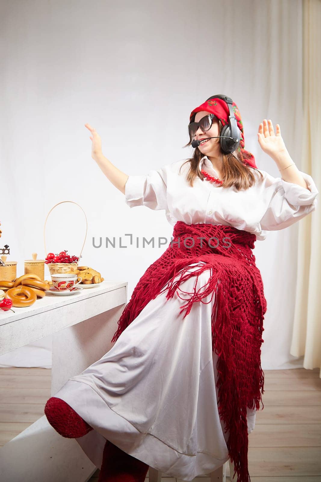 Girl in red scarf and large headphones with microphone having fun at table with delicious food for Orthodox holiday Maslenitsa or Easter. Funny Woman operator, blogger, Freelancer singing and dancing by keleny