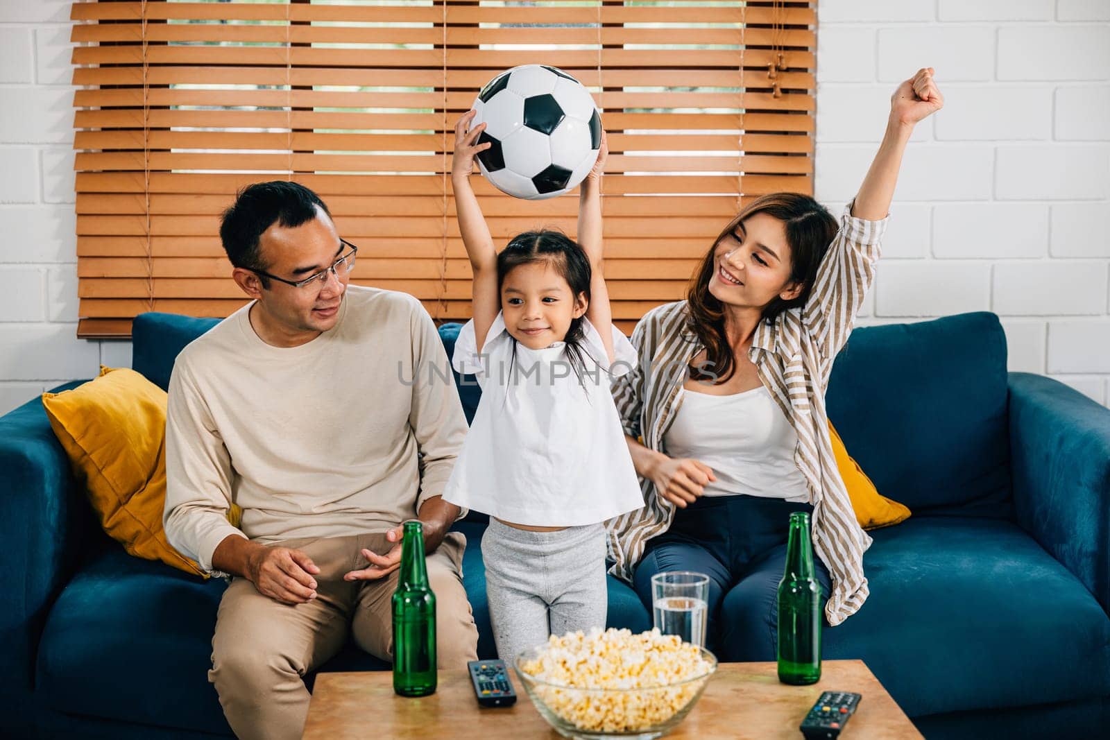 A jubilant family, with popcorn and a ball, raises their arms, cheering while watching a football match at home. Their togetherness and excitement reflect the joy of winning.