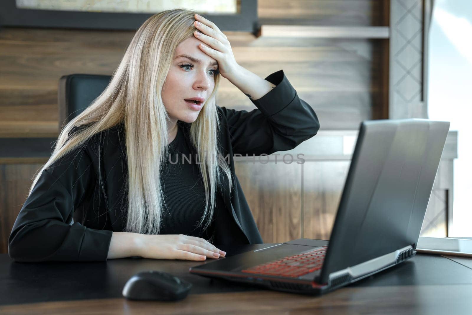 Woman with frightened expression looks at laptop screen, clutching her forehead with her hand in surprise