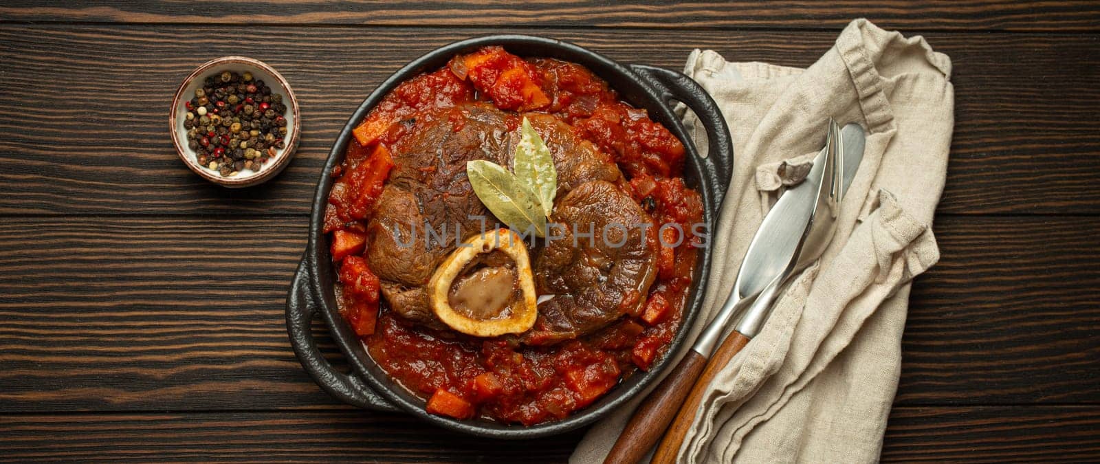 Traditional Italian dish Ossobuco all Milanese made with cut veal shank meat with vegetable tomato sauce served in black casserole pan top view on rustic brown wooden background by its_al_dente