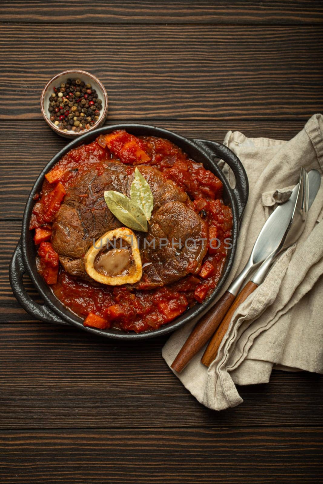 Traditional Italian dish Ossobuco all Milanese made with cut veal shank meat with vegetable tomato sauce served in black casserole pan top view on rustic brown wooden background.