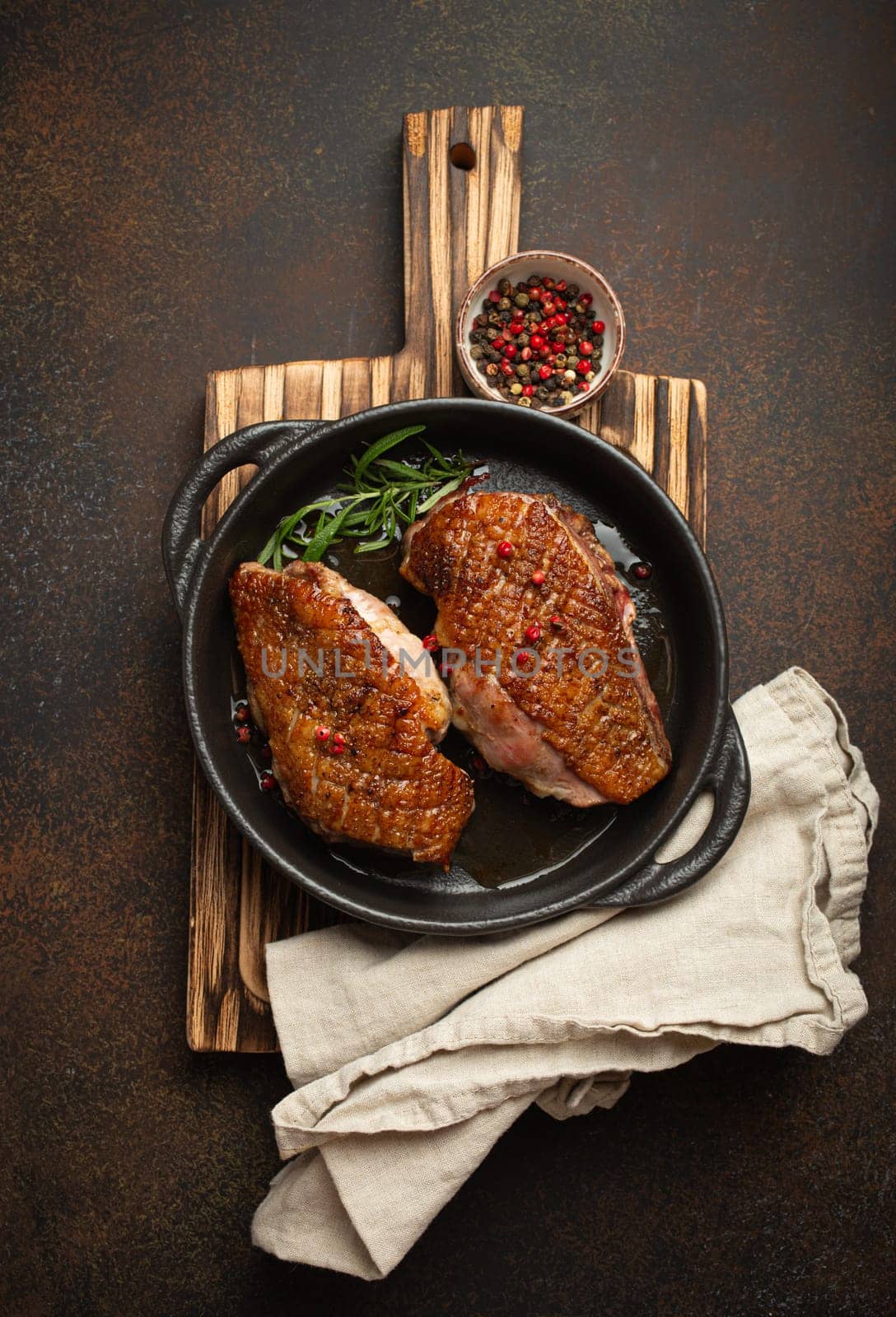 Two roasted duck breast fillets with crispy skin, with pepper and rosemary, top view in black cast iron pan with knife, dark brown concrete rustic background.